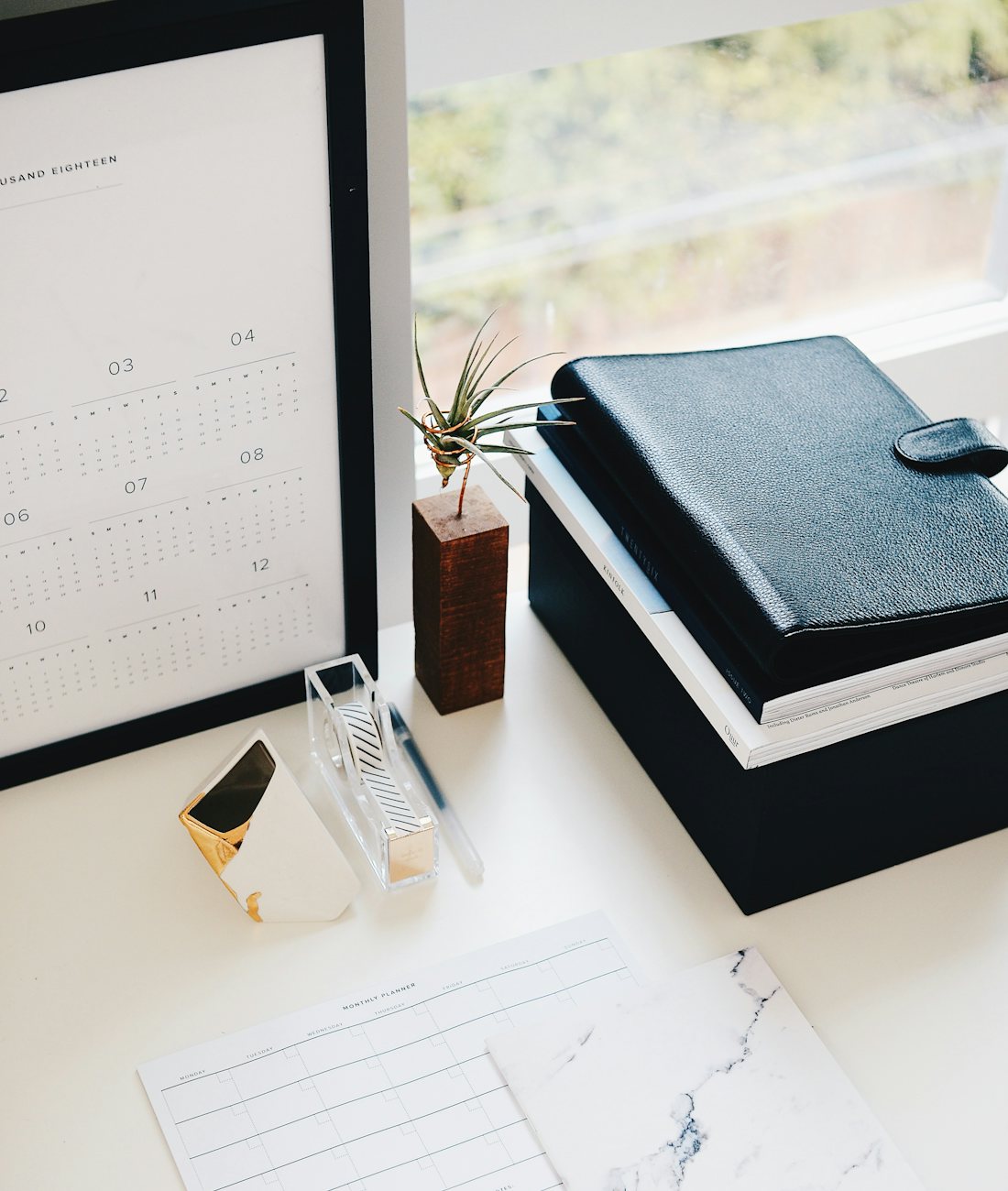 A tidy desk by a window with a year calendar, a leather ledger and a blank monthly planner